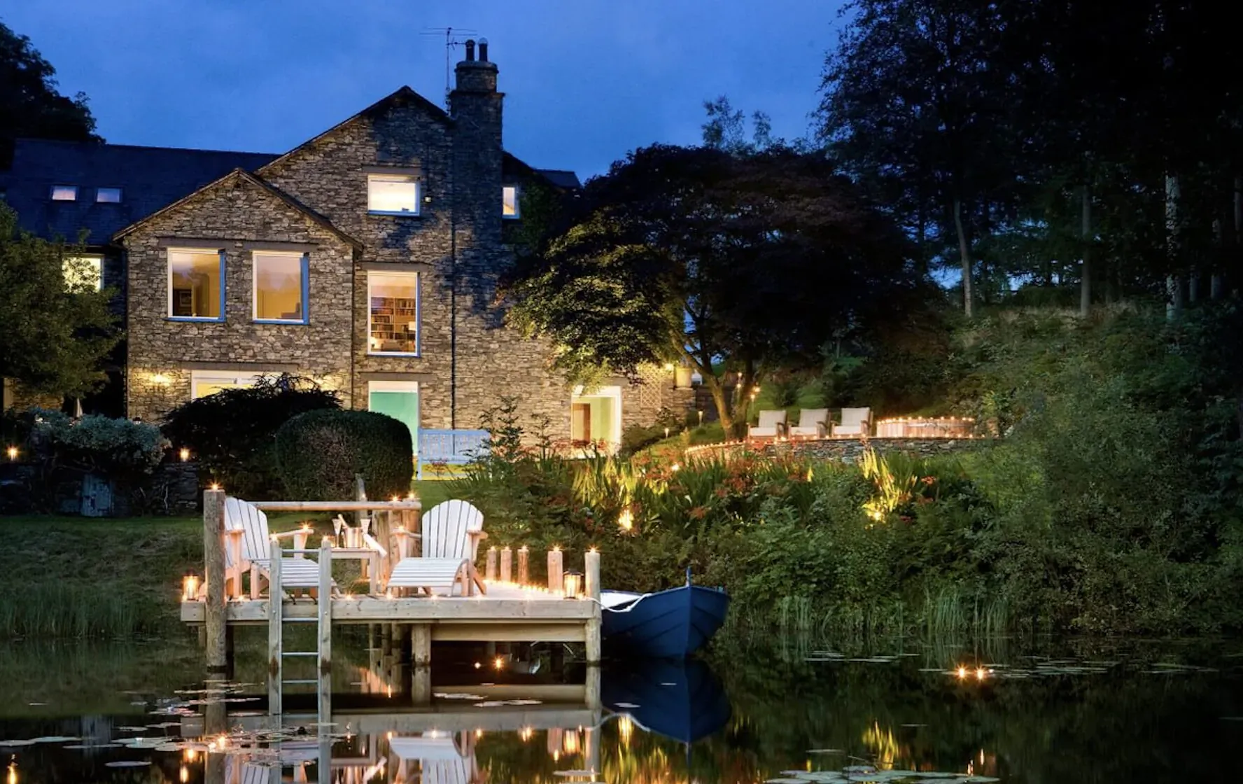 Twilight view of stone luxury house by lake, white chairs on lit wooden dock with rowboat, trees and gardens.