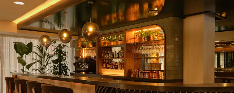 Elegant modern bar at The Reach at Piccadilly, Manchester, with golden pendant lights, plants, bottles, and stools.