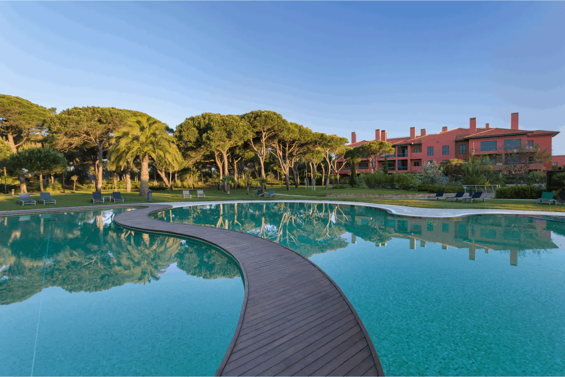Wooden walkway curving through turquoise pool reflecting red Sheraton Cascais Resort buildings and pine trees under blue sky.