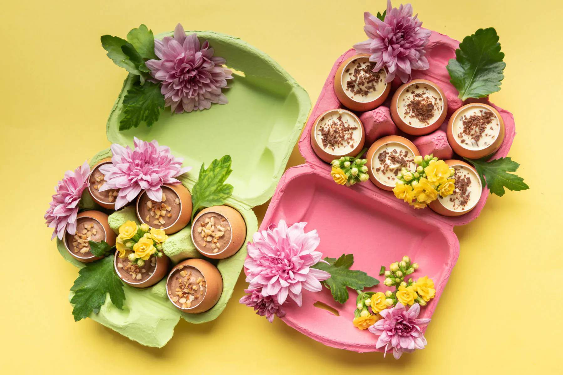 Easter egg-shaped treats topped with cream in green, pink trays garnished with pink dahlias and yellow flowers on yellow background