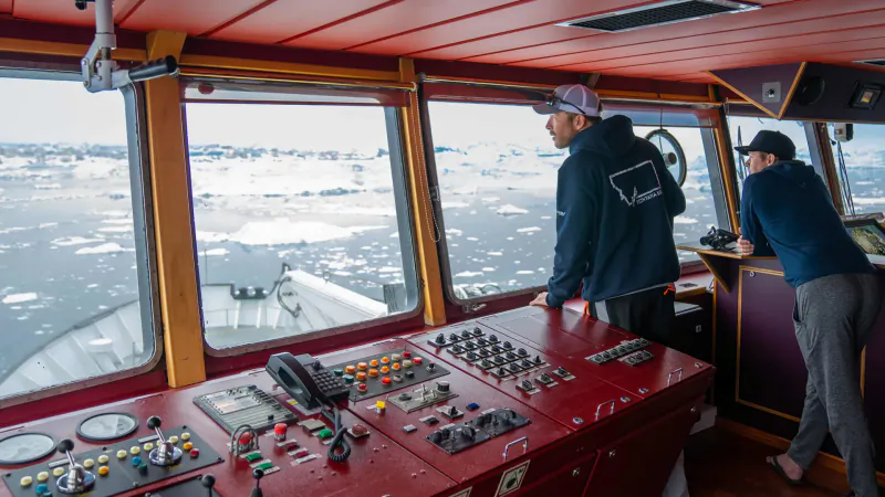 Two crew members at ship's bridge helm, overlooking icy ocean waters through large windows.
