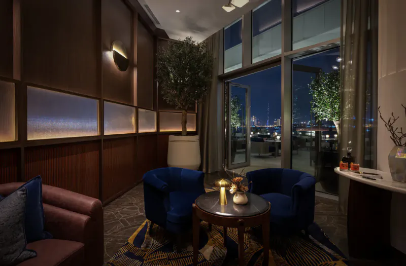 Elegant lounge at Address Grand Creek Harbour with blue velvet chairs, round table, plants, bar, and Dubai night skyline view.