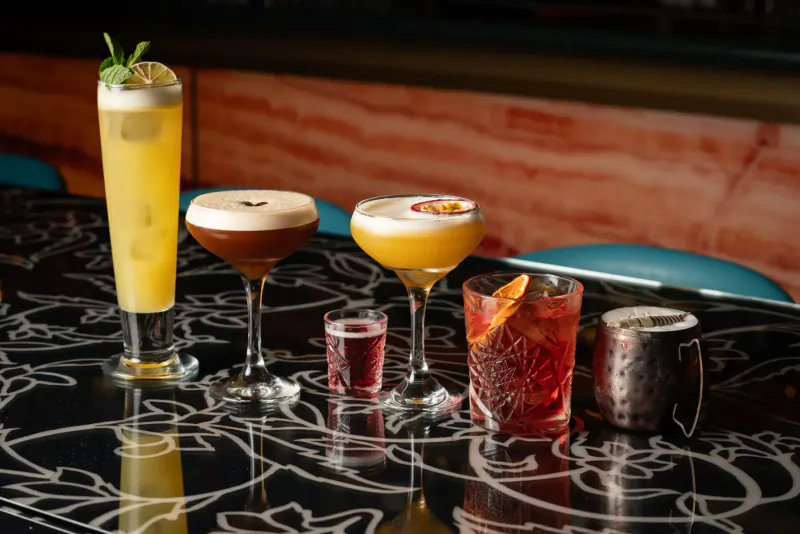 Assortment of colorful cocktails on black bar table: yellow fizz with mint, brown foam, orange twist, and more.
