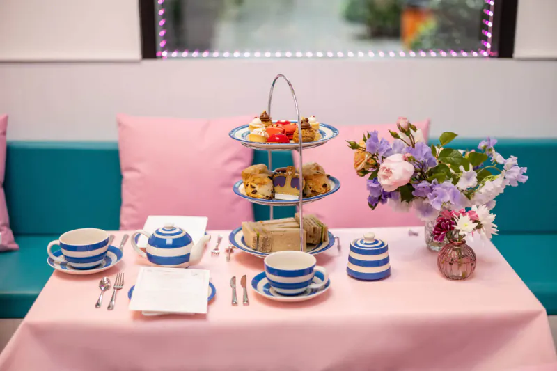 Elegant afternoon tea setup on pink tablecloth with three-tier stand of scones, sandwiches, cakes, teacups, and purple flowers by window.