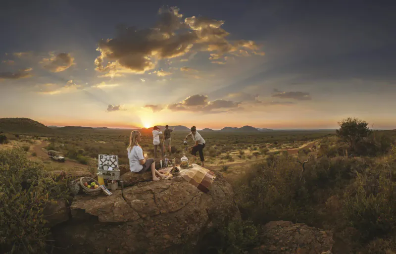 Group of people picnicking on rock outcrop at sunset in Madikwe Game Reserve savanna, Tuningi Safari Lodge.