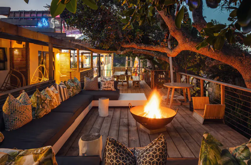 Cozy seaside bungalow deck at dusk with fire pit, L-shaped sofa, chairs under large tree, ocean view.
