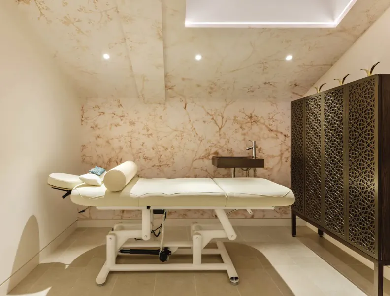 Elegant spa treatment room with white massage table, beige marble walls, recessed lighting, sink, and ornate cabinet.