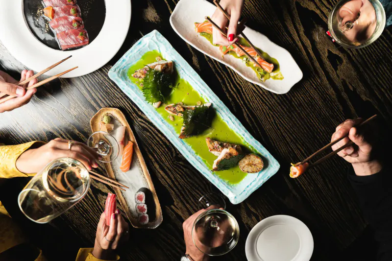 Top-down view of sushi, sashimi, and green sauce dishes on table, hands using chopsticks and wine glasses at Nobu festive meal