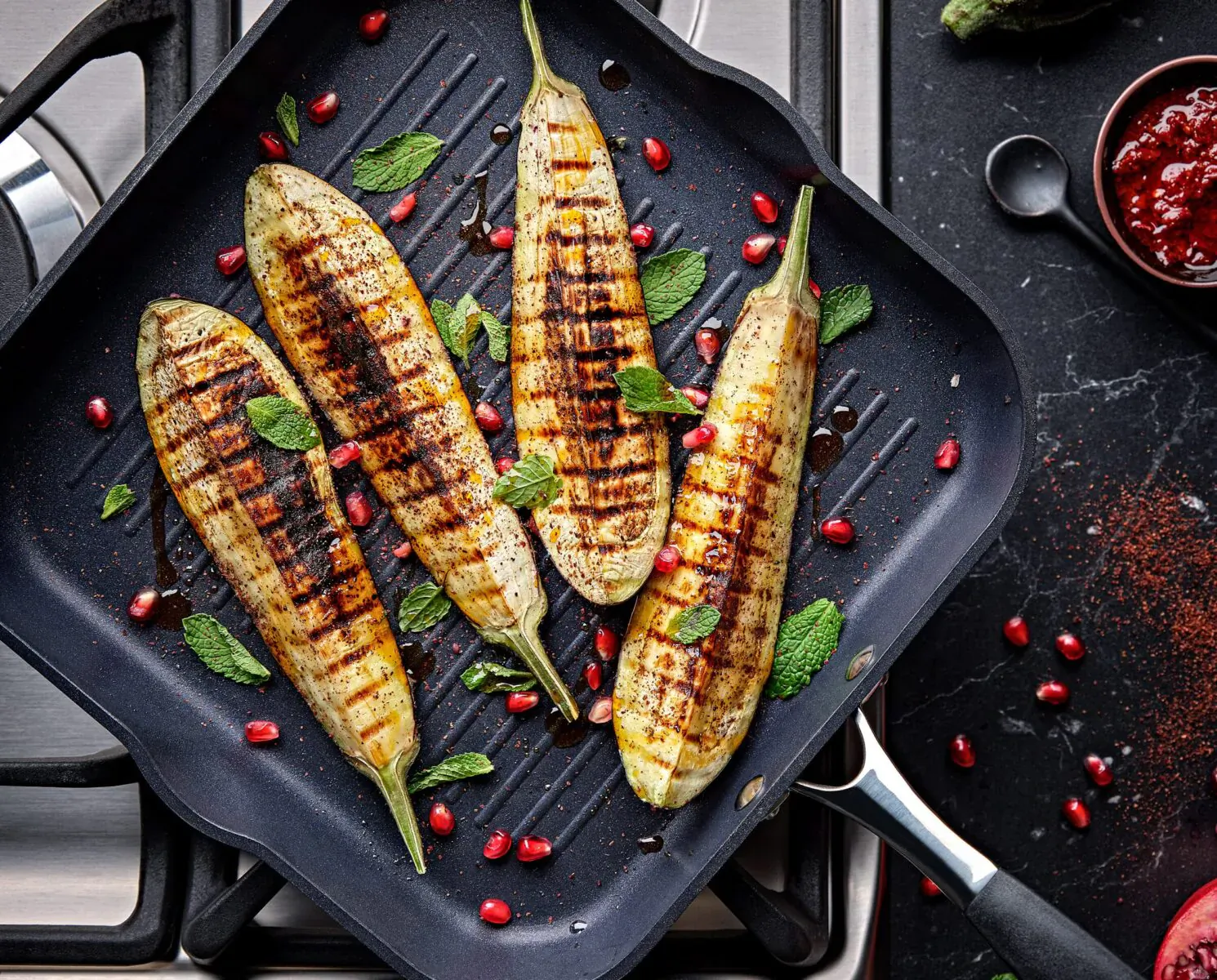 Grilled eggplants with pomegranate seeds and mint on a black square grill pan on stovetop.