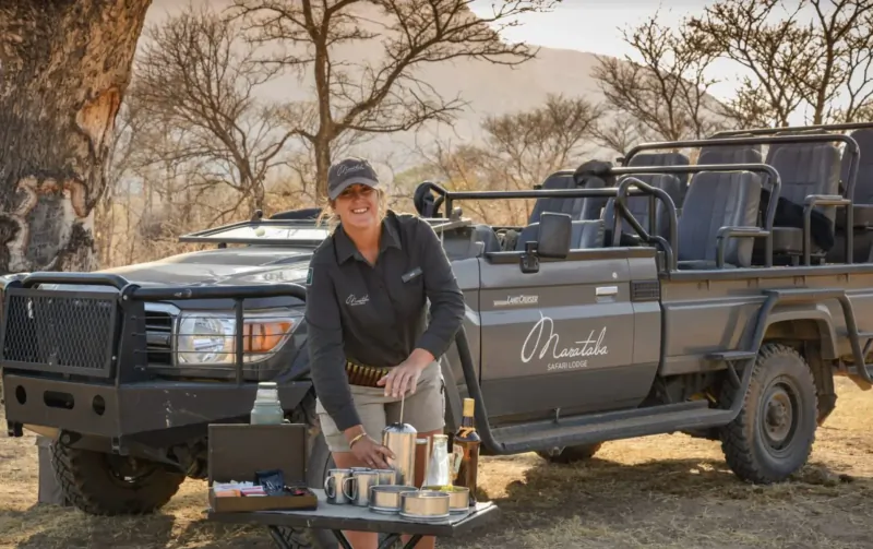 Smiling woman in cap and khaki leans on Marataba safari vehicle with drinks on table amid African bushveld.