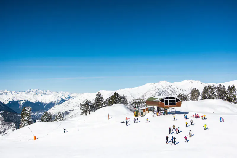 Skiers and snowboarders on snowy slopes in Val d'Aran Pyrenees, with modern lift station and distant snow-capped mountains under blue sky.