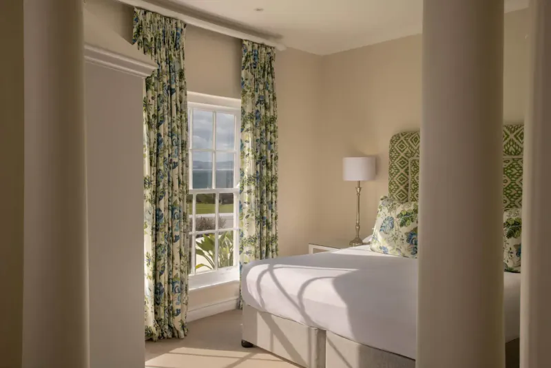 Elegant hotel bedroom with floral green curtains, white bed, lamp, and mountain view through window.