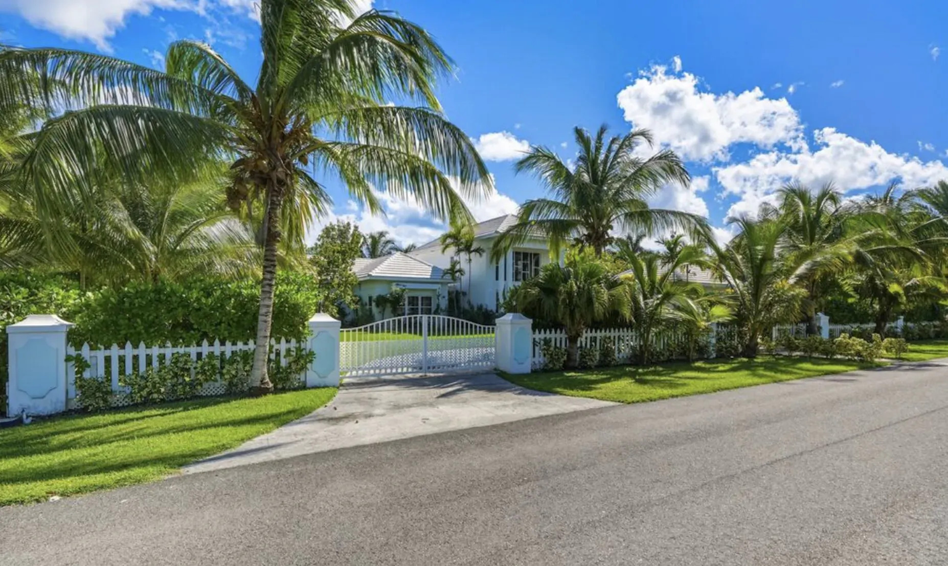 Ciao Bella luxury estate in Lyford Cay, Bahamas: white house behind gated white picket fence, palm trees, lush lawn, blue sky.