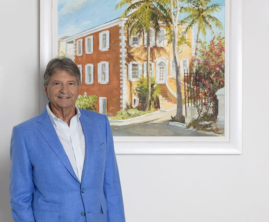 George Damianos, CEO of Bahamas Sotheby’s, in blue suit, smiling beside painting of tropical Bahamian house with palms.