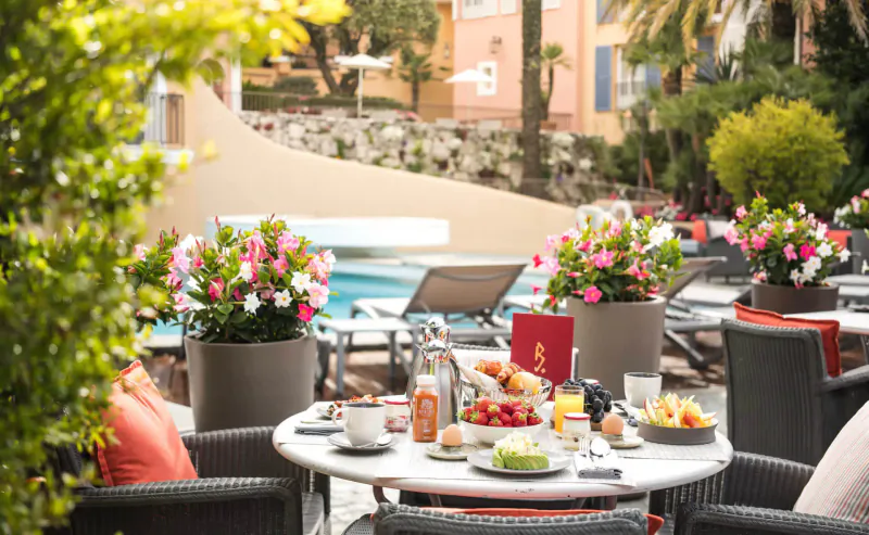 Luxury outdoor breakfast table with fruits, coffee, juices amid vibrant flowers, pool, and ochre buildings at Byblos Palace hotel, Saint-Tropez