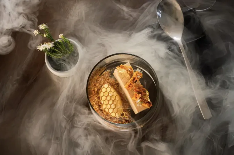 Glass dish of honeycomb, white slab, and sauce with steam rising, spoon, daisies on dark table