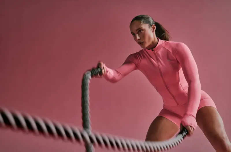 Athletic woman in pink zip-up bodysuit gripping thick battle ropes, poised to train against pink background