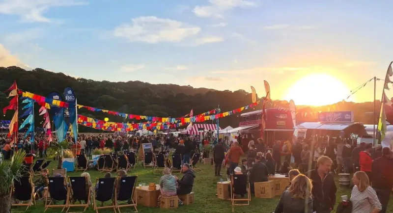 Crowded Travelling Feast Festival at sunset with colorful flags, tents, people sitting and standing on grass amid hills.