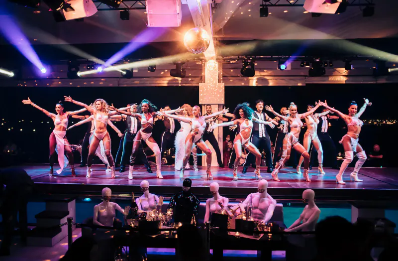Lio Cabaret Ibiza: Dancers in glamorous outfits perform energetically on stage with live band, vibrant pink lighting.