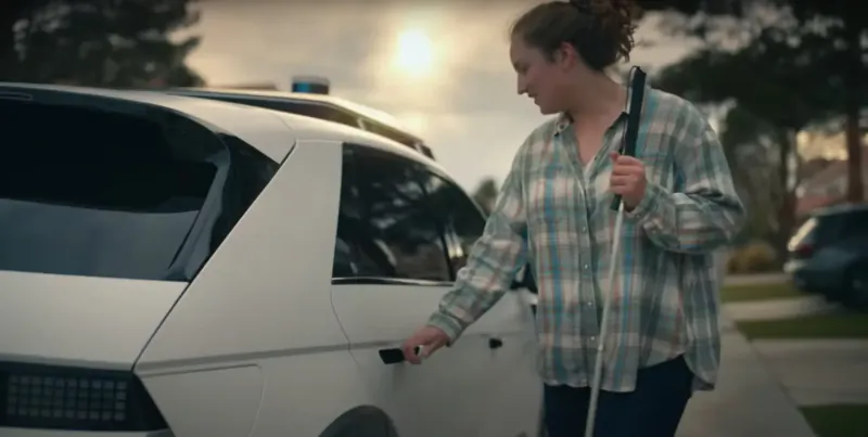 Woman with white cane opening door of white Hyundai IONIQ 5 robotaxi with police lights at sunny suburban driveway