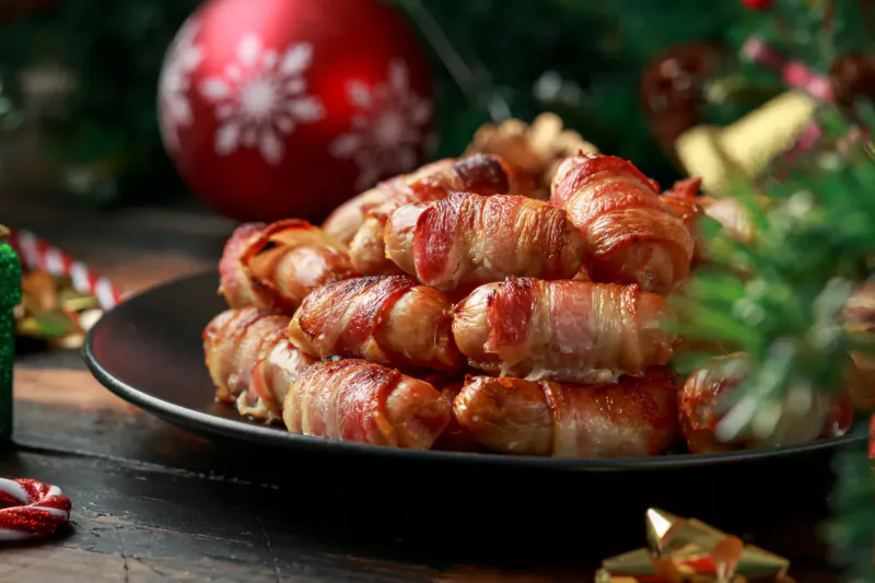 Stack of bacon-wrapped sausages on black plate amid Christmas decorations and ornaments