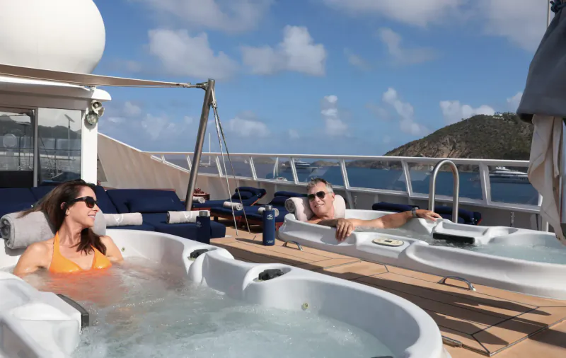 Couple relaxing in adjacent hot tubs on luxury yacht deck, sunny ocean view with hills.