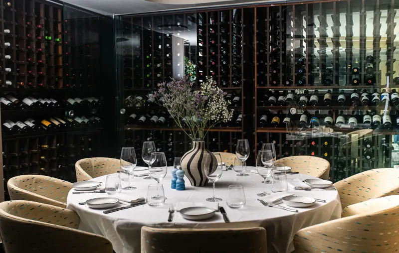 Elegant round table set with white linens, wine glasses, blue-striped vase of lavender flowers, salt/pepper shakers in wine cellar with racks.