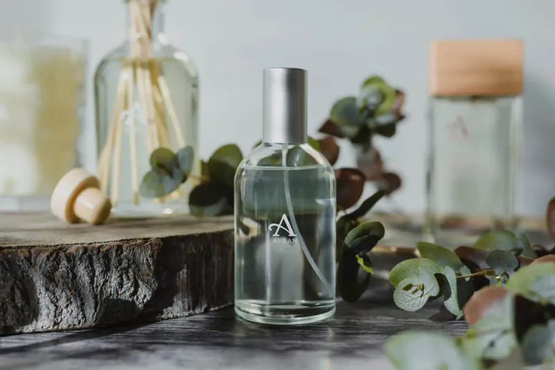 ARRAN Sense of Scotland clear perfume bottle with 'A' label, surrounded by eucalyptus leaves, reed diffuser, candle on wood base