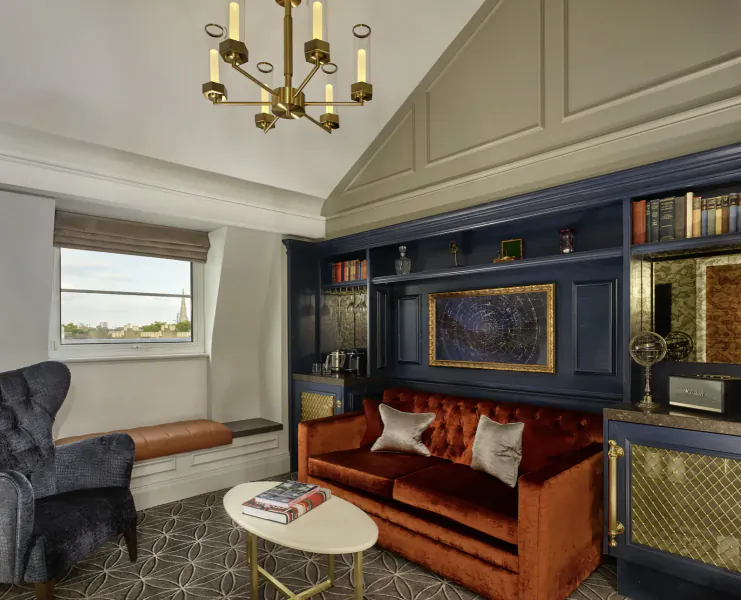 Elegant hotel lounge with tufted orange sofa, blue bookcase, chandelier, and window in luxury Gallery room at 100 Queen's Gate Hotel.