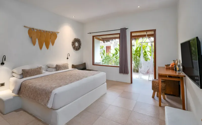 Spacious white bedroom at Amber Lombok Beach Resort with king bed, desk, TV, leaf wall decor, and open doors to tropical garden.