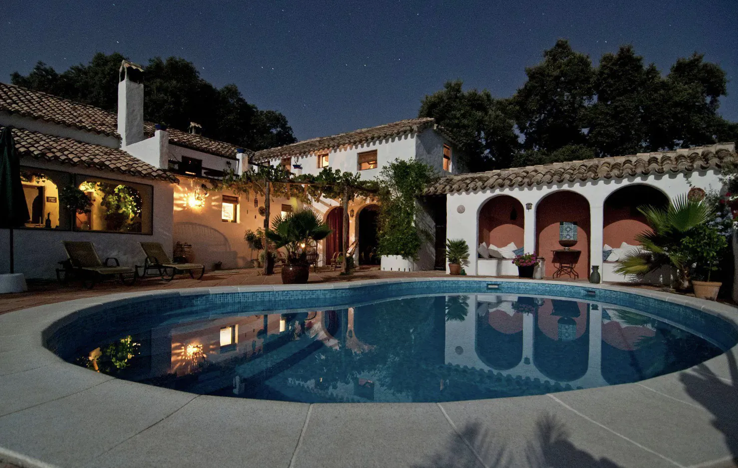 Night view of luxurious Spanish villa courtyard with lit oval pool, lounge chairs, and palm trees.