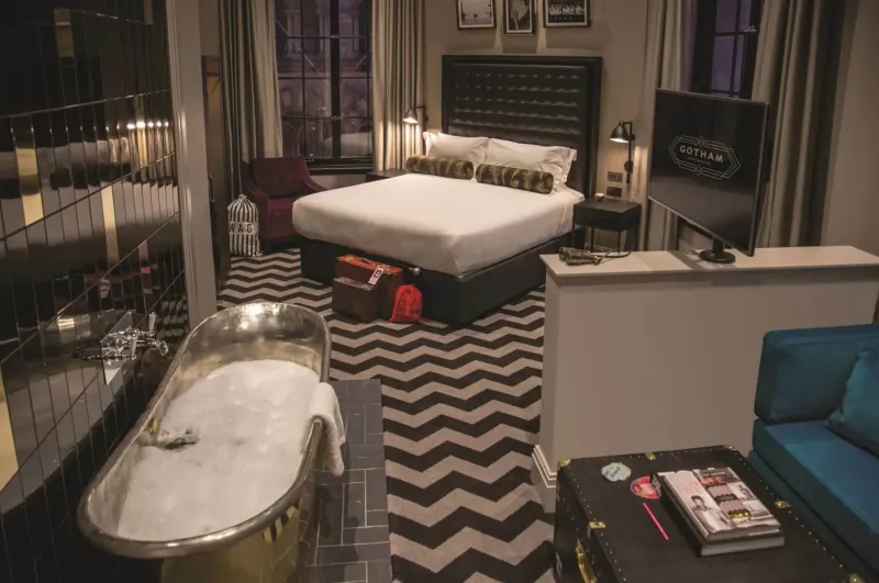 Luxurious Hotel Gotham suite with black-and-white zig-zag carpet, king bed, clawfoot bathtub, vintage suitcases, and TV.