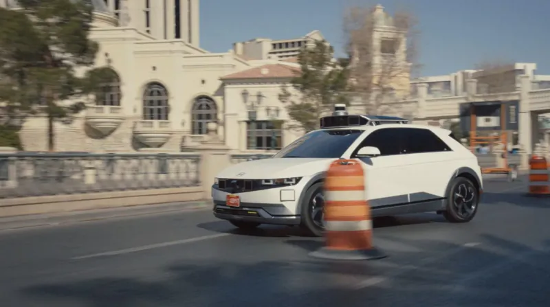 White Hyundai IONIQ 5 robotaxi with lidar drives past orange traffic cones near DMV building during driving test.