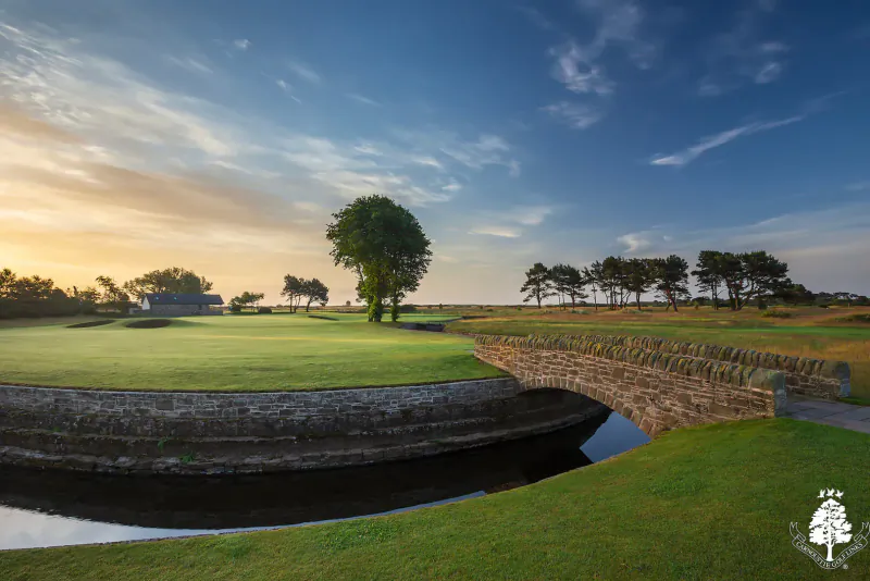 Carnoustie Links golf course at sunset: curved green bunker with stone bridge over stream, trees, and clubhouse.