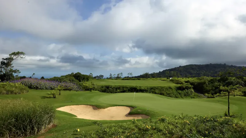 Heart-shaped sand bunker on lush green golf course at Avalon Golf Estate, Mauritius, with hills and clouds.
