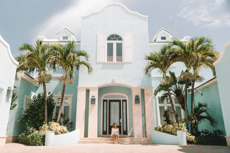 Woman sits on steps of dreamy light blue tropical mansion with palm trees and gardens