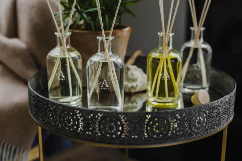 Four Arran reed diffuser bottles in clear glass with yellow oils on ornate black tray, plant beside.
