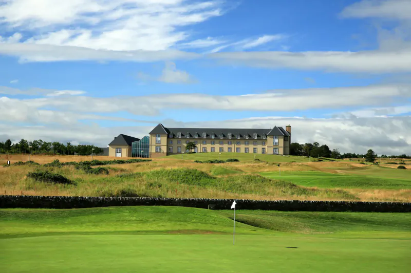 Large beige stone building on grassy golf course hills under partly cloudy blue sky, with flagstick in foreground.