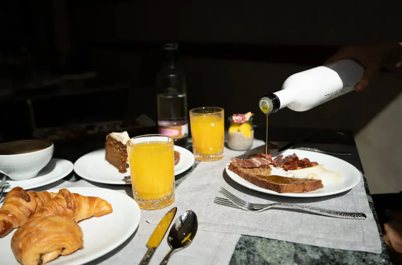 Hand pouring honey from white jar onto croissant on breakfast plate with eggs, cake, orange juice, and coffee.