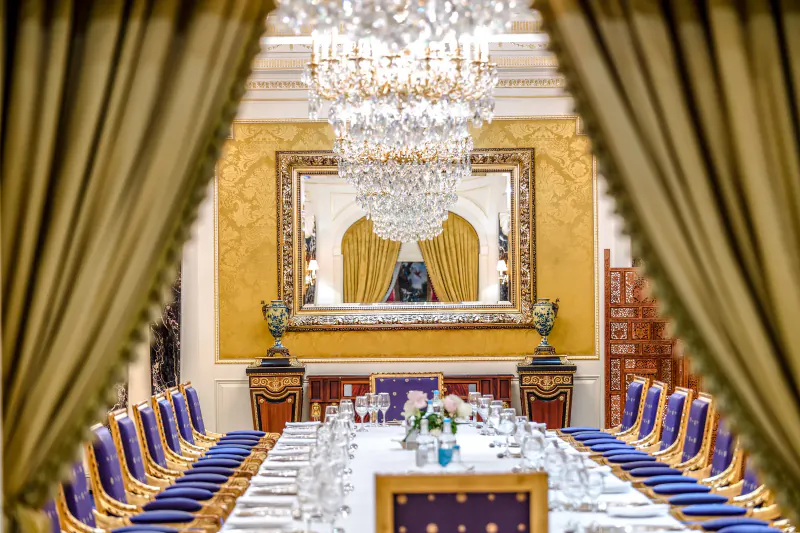 Elegant dining room at Bentley Hotel with crystal chandelier, gold-framed mirror, blue velvet chairs, and white-draped table in opulent setting.