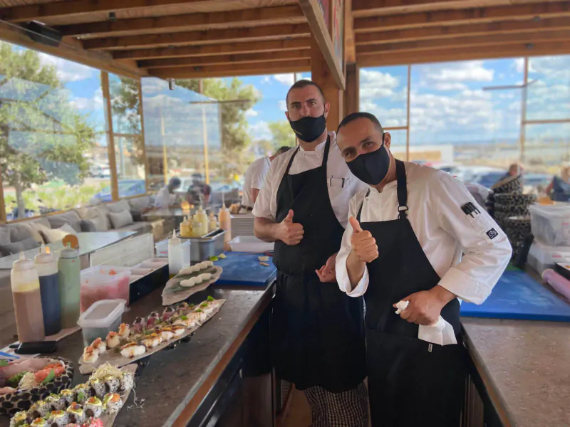 Two masked chefs in white uniforms give thumbs up behind sushi and seafood buffet at Assaona Gastrobeach Palma Club.
