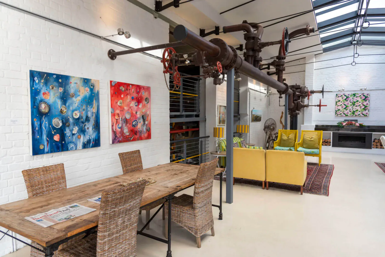 Industrial chic hotel interior with exposed pipes, abstract art, wooden dining table, yellow sofas, and fireplace.