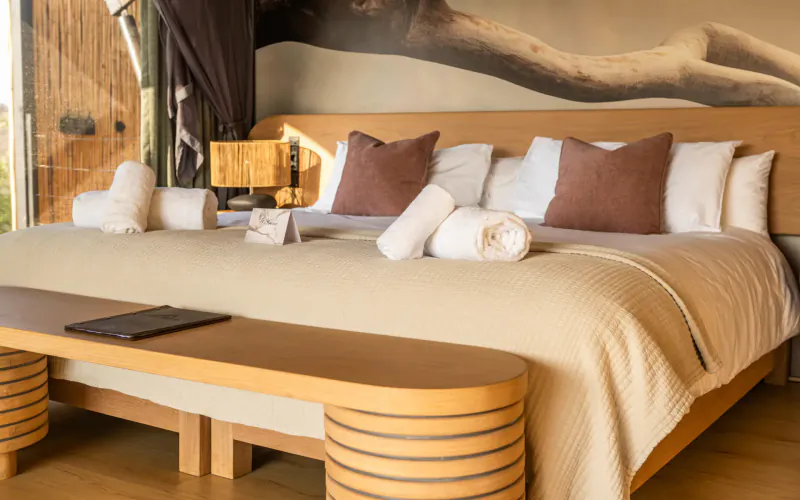 Luxurious king bed in 18 On The Hill suite with wooden headboard, rolled towels, beige duvet, and tree mural backdrop.