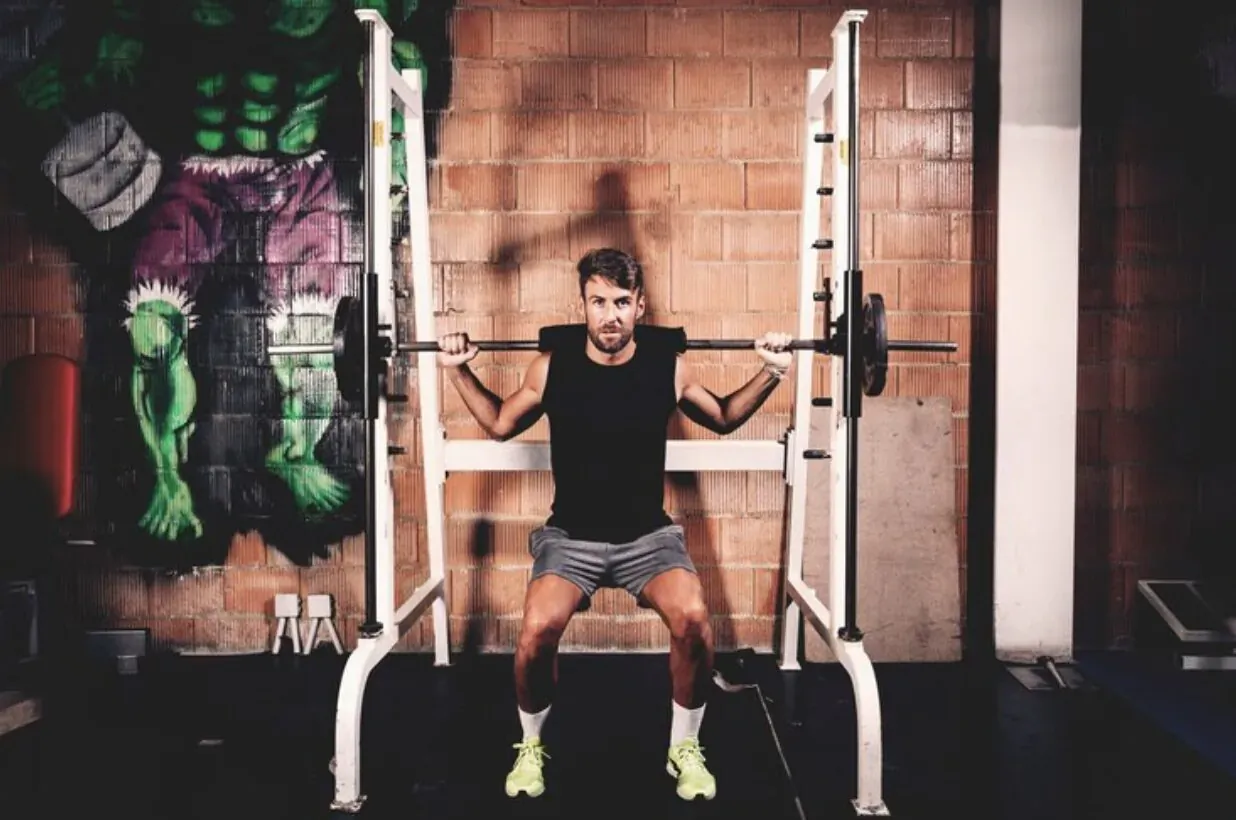 Man squatting with barbell in power cage, Hulk mural on brick gym wall