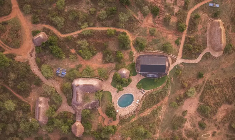 Aerial view of luxury eco-resort with thatched huts, solar-roofed building, pool, and dirt paths in savanna landscape