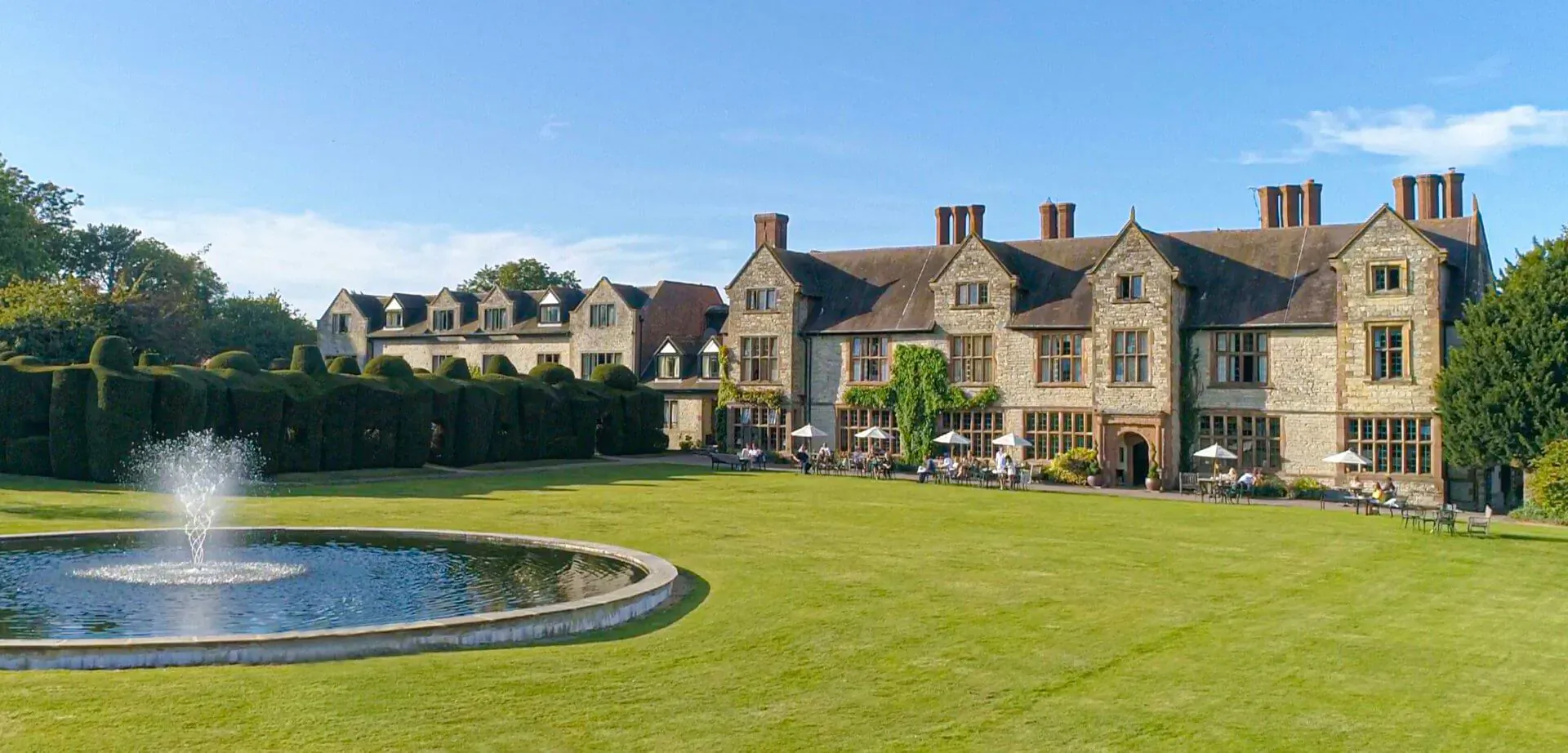 Billesley Manor Hotel & Spa, historic stone manor with chimneys, fountain, lawns, and gardens under blue sky