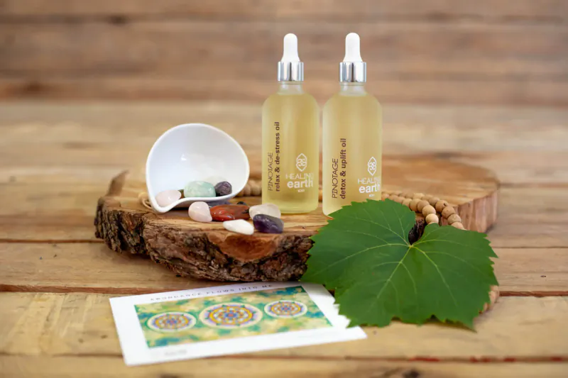 Two 'Healing Earth' oil dropper bottles on wooden base with crystals, green leaf, and mandala card