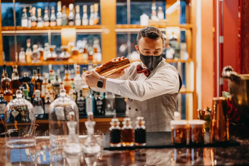 Bartender in bow tie and mask shaking cocktail at upscale bar with extensive liquor shelves and glassware.