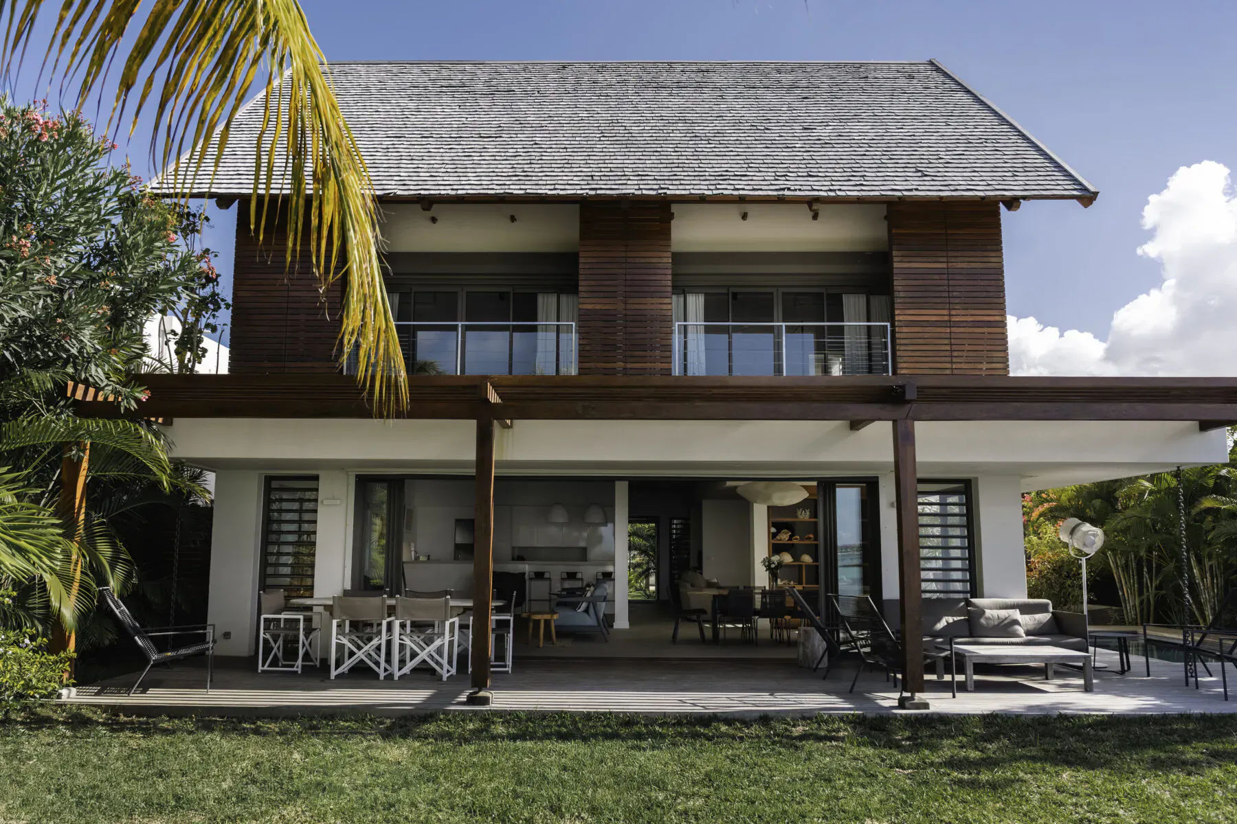 Modern tropical villa in Mauritius with wooden facade, spacious veranda, outdoor dining, and lush garden under blue sky.