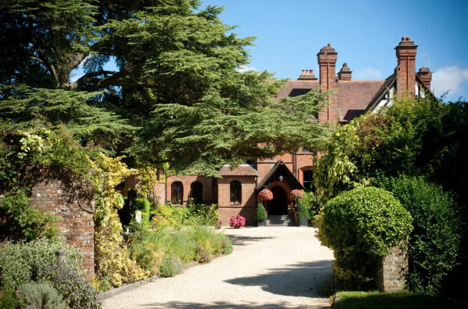 Careys Manor Hotel & SenSpa, luxurious red-brick manor with arched entrance, surrounded by lush green gardens and trees under blue sky.